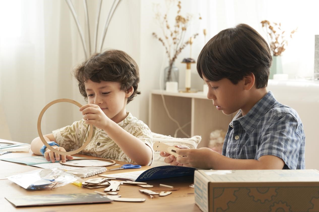 Two young boys are engrossed in a construction project at a table. The boy on the left, with curly hair and a patterned shirt, examines a circular wooden piece, while the boy on the right, wearing a blue checkered shirt, carefully handles a smaller wooden component. Scattered around them are instruction booklets and various other wooden parts, suggesting they are building a model or a STEM-related toy from a kit.