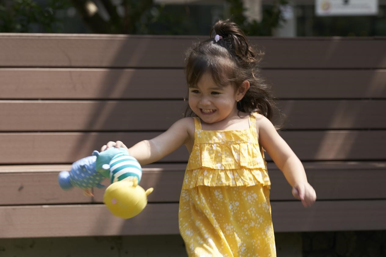 A young girl in a sunny park runs towards the camera with a joyful expression. She wears a bright yellow dress and her dark hair is pulled back in a high ponytail. The girl carries a plush toy with her, swinging it playfully as she moves.