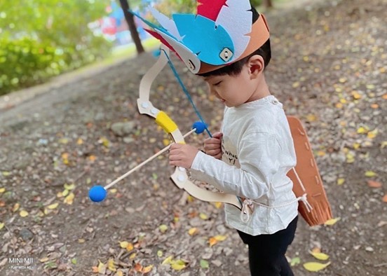 A young boy, dressed in a white shirt and black pants, is holding a toy bow and arrow. He is wearing a colorful feathered headdress and has a serious expression on his face as he aims. The scene is playful and imaginative, capturing the joy of childhood and the power of pretend play.