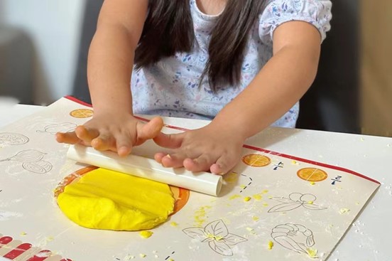 A young girl is rolling out a yellow piece of playdough on a white surface. She is using a white rolling pin and her hands are covered in flour. The scene is playful and fun, capturing the joy of childhood and the creativity of play.