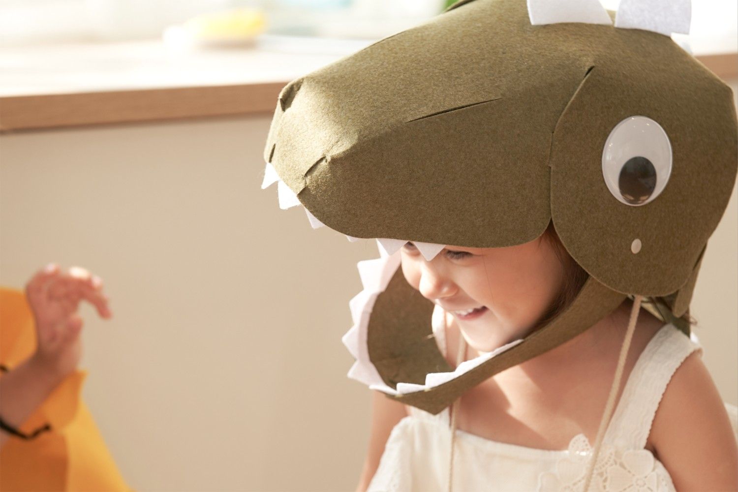 A young girl, wearing a felt dinosaur hat, smiles brightly as she looks down. The hat is a light brown with a white underbite and a large, black eye. The girl is wearing a white dress with a lace trim and her hair is pulled back. She is sitting in a room with a white wall and a wooden shelf in the background. The scene is full of childlike wonder and the joy of play. It looks like the girl is having a lot of fun pretending to be a dinosaur.