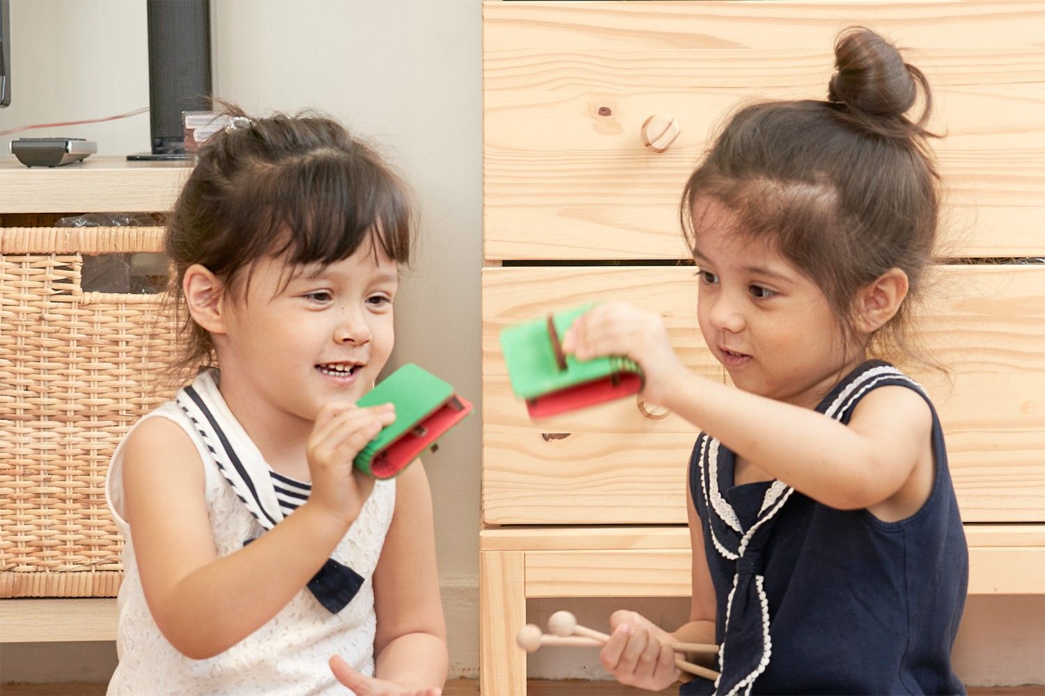 Two young girls are playing with musical instruments. The girl on the left is holding a pair of wooden clappers, and she is smiling happily. The girl on the right is holding a pair of wooden drumsticks, and she is looking at her friend with a mischievous grin. The girls are both wearing sailor-style tops, and they look like they are having a lot of fun making music together. The scene is full of childlike joy and the simple pleasures of play. It looks like the girls are about to create a beautiful melody.