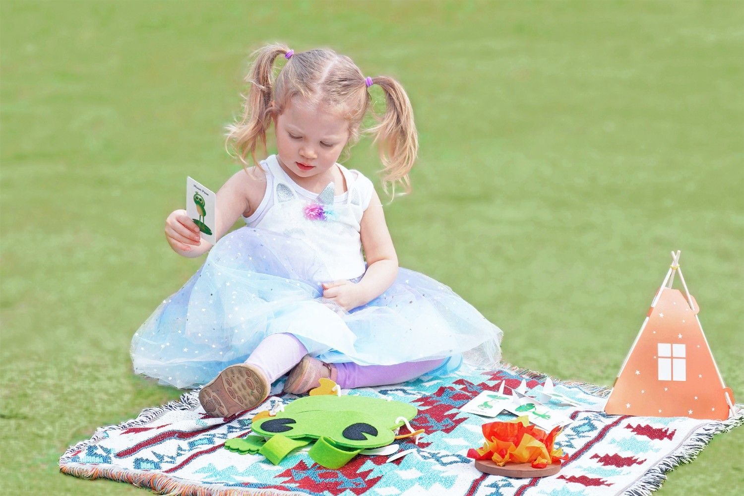 A young girl, wearing a blue tulle dress and her hair in pigtails, is sitting on a colourful rug in a grassy field. She is holding a card with a picture of a frog on it, and she is looking at it with a thoughtful expression. There is a small orange tent and a green frog puppet on the rug. The scene is full of childlike wonder and the joy of play. It looks like the girl is enjoying a quiet moment of imaginative fun, and she is using her imagination to create a world of her own.