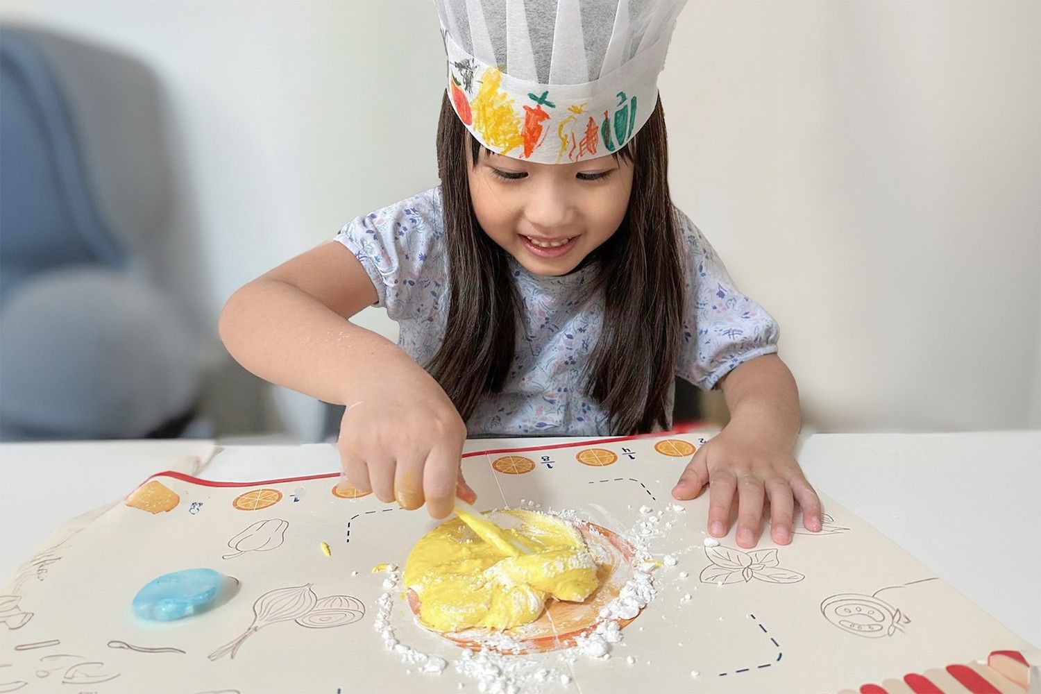 A young girl, wearing a white chef's hat decorated with colourful vegetables, is smiling as she uses a yellow plastic tool to spread a yellow substance on a table. There is white powder sprinkled around the yellow substance and a blue object to the left. The table is covered with a white tablecloth with a red and white striped border. The girl is clearly enjoying her culinary adventure, and she looks like she is about to create something delicious. It is a heartwarming scene of a child's imagination and creativity.