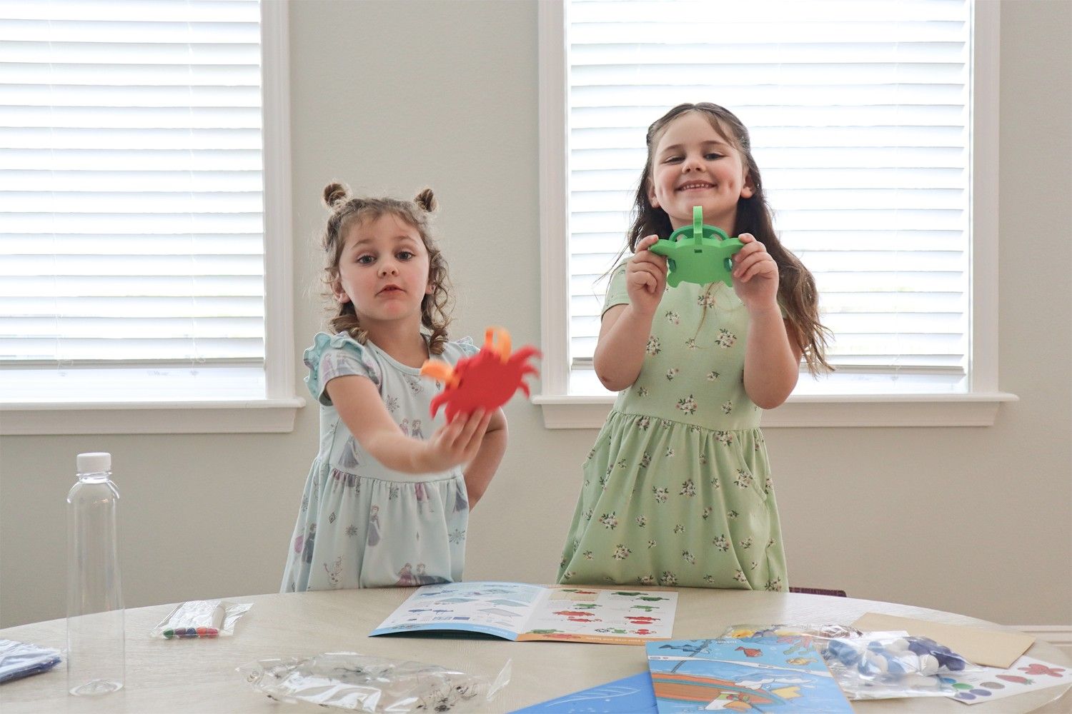 Two young girls are sitting at a table, each holding a colourful sea creature toy. The girl on the left is wearing a blue and white dress and holding a red crab. The girl on the right is wearing a green floral dress and holding a green crab. Both girls are smiling and looking at the camera. On the table, there are some craft supplies, including a bottle of water, some markers, and a booklet of instructions. The scene is full of childlike joy and the simple pleasures of play. It looks like the girls are having a lot of fun learning about sea creatures and making crafts together.