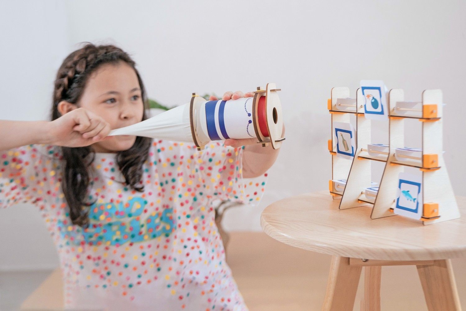 A young girl, wearing a white and colourful polka dot top, is using a paper cone to blow air into a small wooden device. The device is attached to a wooden stand with a red spool of string. There are three cards on the stand, each with a different picture of a sea creature. The girl is concentrating hard, and she looks like she is enjoying her experiment. The scene is full of childlike curiosity and the joy of discovery. It looks like the girl is about to learn something new and exciting about the world around her.