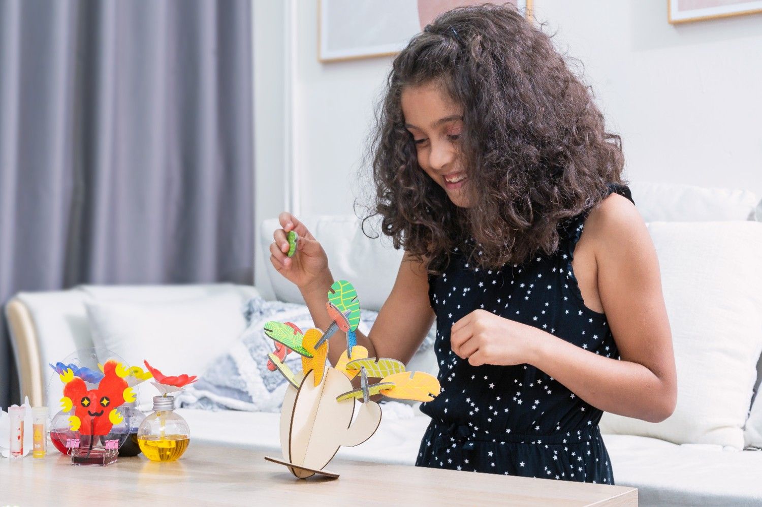 A young girl with long, curly brown hair, wearing a black and white polka dot dress, is smiling as she carefully places a green leaf onto a wooden cactus model. The cactus is part of a science experiment, and there are two small glass bottles filled with colourful liquids nearby. The girl is clearly enjoying herself, and she is excited to see how the experiment will turn out. The scene is full of childlike curiosity and the joy of learning. It looks like the girl is having a lot of fun exploring the world around her.