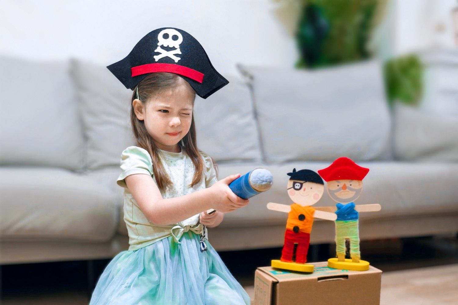 A young girl, dressed as a pirate with a black felt hat adorned with a skull and crossbones, is holding a blue toy sword. She is winking at the camera and looking determined. The girl is sitting on the floor in front of a grey sofa, and there are two wooden pirate figures standing on a cardboard box behind her. The scene is full of childlike fun and imagination. It looks like the girl is ready for adventure, and she is using her imagination to create a world of her own.