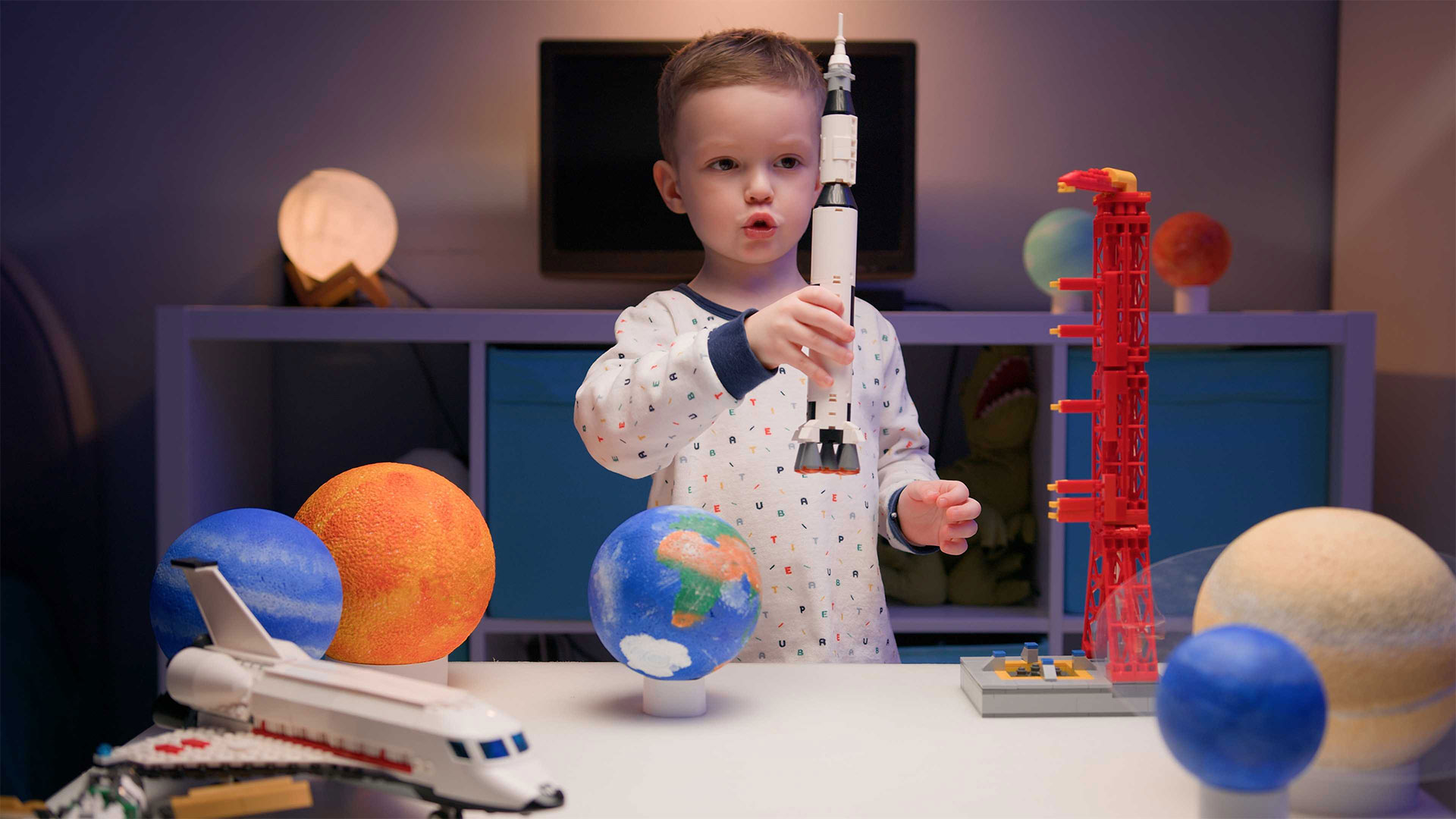 A young boy, wearing a white and blue pajama top, is playing with a toy rocket and other space-themed toys, engaging in hands-on STEM education about space exploration. He is holding the rocket up in the air, and he has a look of wonder on his face. There are several other toys on the table, including a model of a space shuttle, a model of a launchpad, and several planets. The scene is full of childlike imagination and the excitement of discovery. It looks like the boy is about to embark on a grand adventure into space.