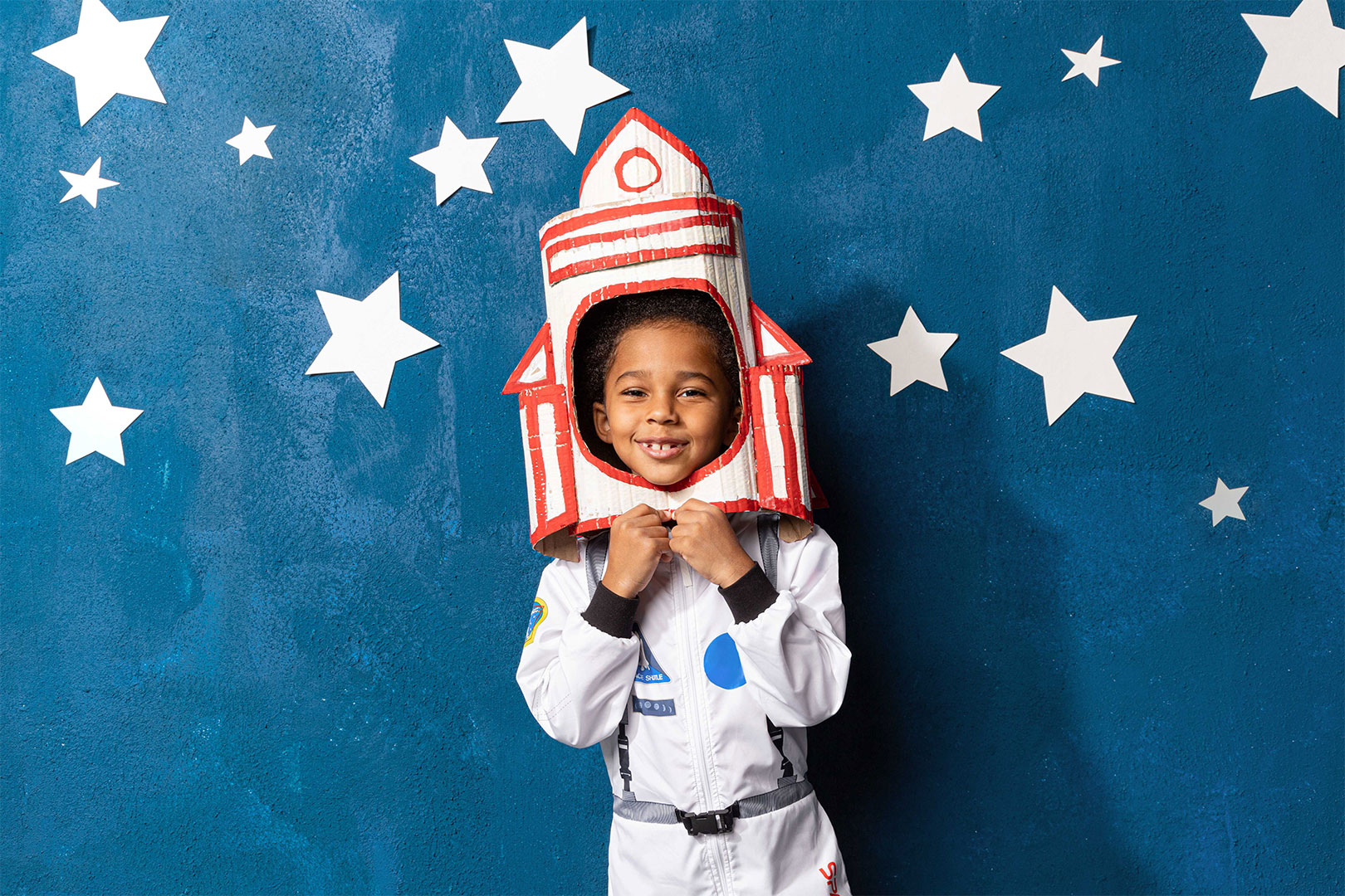 A young boy, dressed in a white astronaut suit, stands in front of a blue wall adorned with white paper stars. He is wearing a cardboard rocket helmet, and he is holding the edges of the helmet with both hands. He is smiling broadly, and his eyes are shining with excitement. The scene is full of childlike wonder and the joy of imagination. It looks like the boy is ready to blast off on an adventure into space.