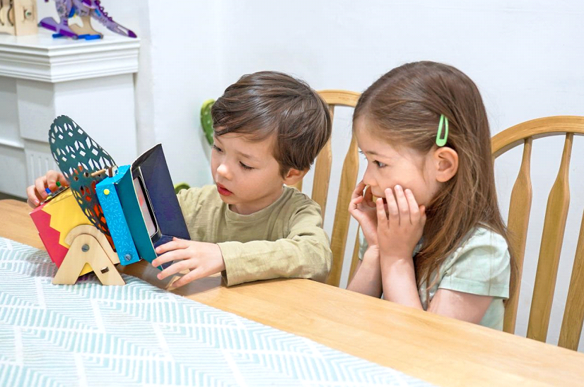 A young boy is sitting at a table, looking intently at a colourful toy that he is holding in his hands. He is wearing a brown t-shirt, and his hair is short and dark. A young girl is sitting next to him, her hands clasped in front of her face. She is wearing a green t-shirt, and her hair is long and brown. The table is covered in a blue and white patterned tablecloth. The scene is full of childlike curiosity and wonder. The boy is clearly engrossed in his toy, and the girl is watching him with interest. It looks like they are about to have a lot of fun together.