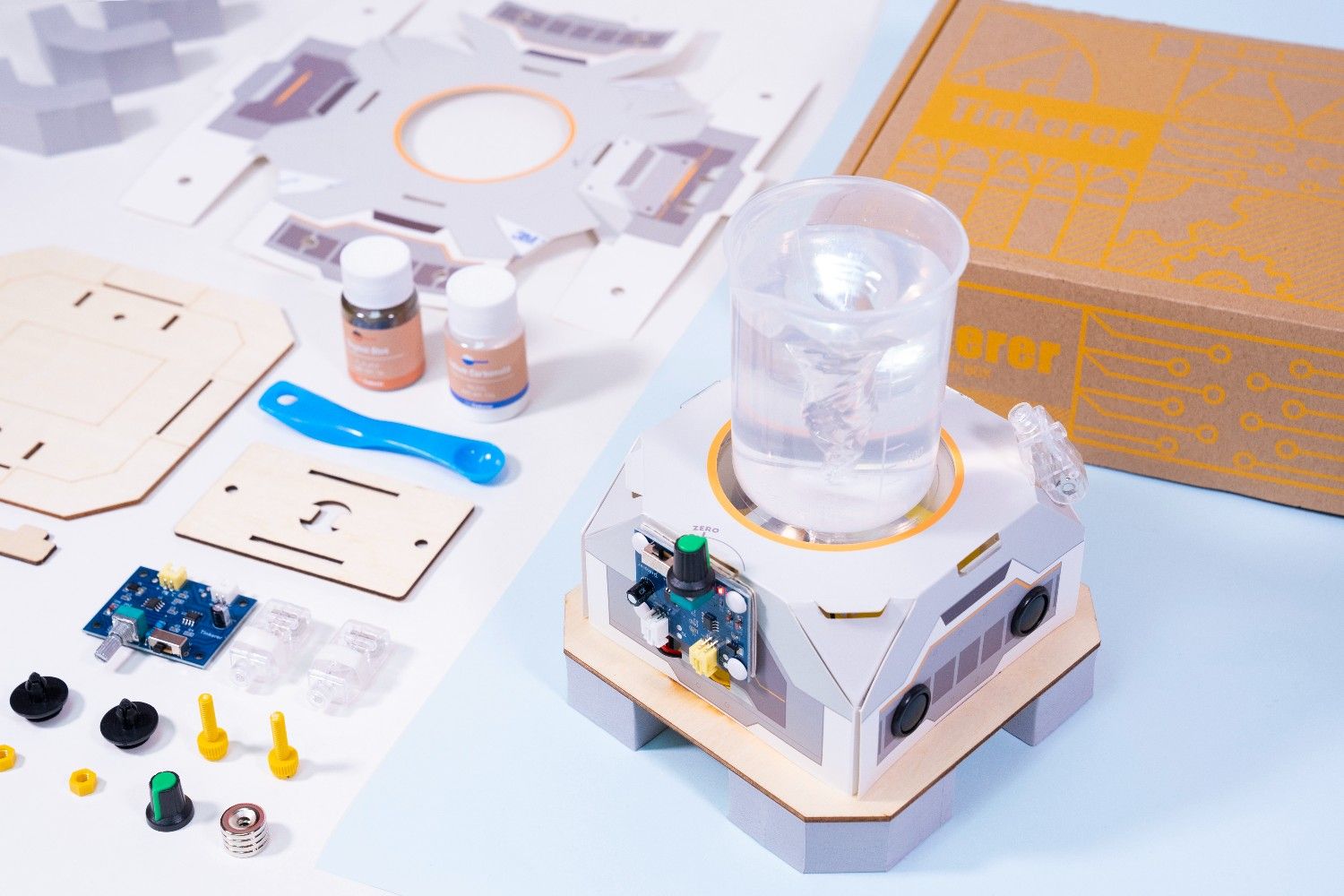 A partially assembled science kit sits on a light blue surface. The kit includes a clear plastic container with a swirling vortex of water, a cardboard box with a circuit board design, a wooden base, a blue plastic tool, and a variety of small parts, including buttons, knobs, and wires. The kit looks like it would be a fun and engaging way to learn about science and engineering. The swirling water is particularly mesmerising, and the kit's overall design is sleek and modern. It looks like a great way to spark a child's curiosity and encourage them to explore the world around them.