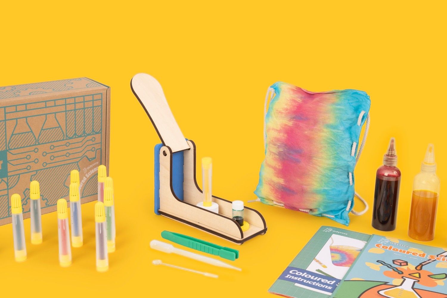 A vibrant collection of tie-dye supplies is spread out on a bright yellow background. There's a cardboard box with a circuit board design on the side, a small wooden tie-dye kit with a lever, a colourful tie-dye bag, two bottles of dye, a booklet of instructions, and a set of colourful markers. The scene is full of creative energy and the promise of fun and colourful creations. It looks like a fun and engaging activity for children, and it is a great way to teach them about art, science, and creativity.