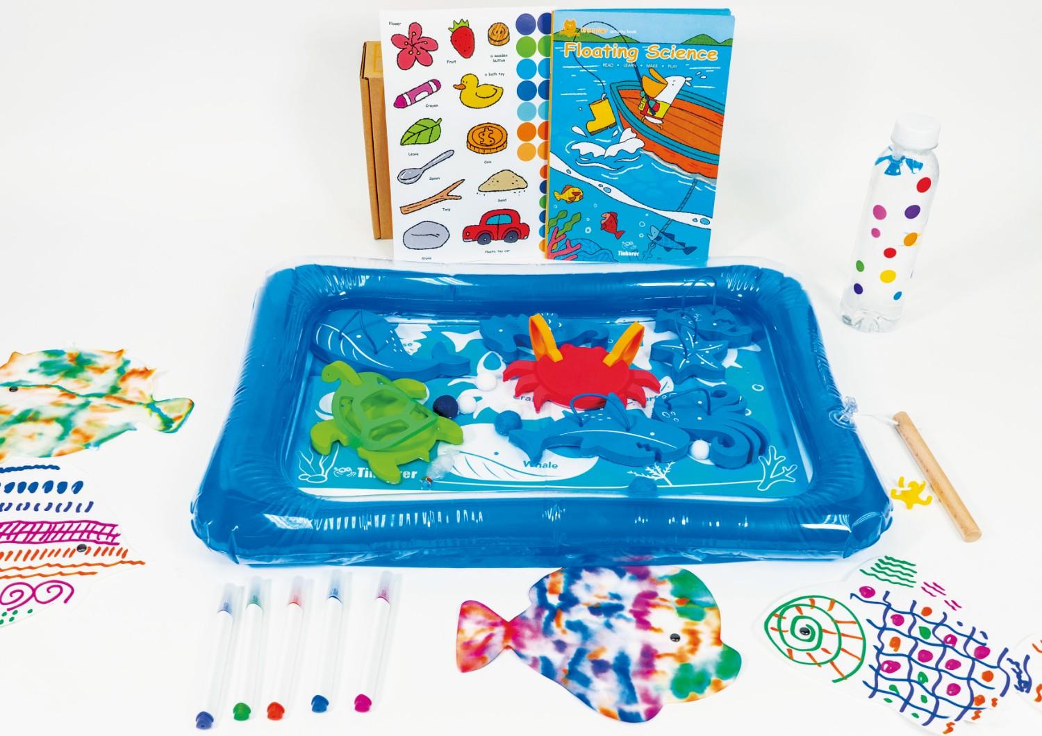 A blue inflatable tub filled with water sits in the centre of the image. The tub is filled with sea creatures, including a red crab, a green turtle, and a blue whale. There are also some white balls floating in the water. A wooden stick is used to pull the objects around the tub. On the left side of the image, there are four colourful markers and three paper fish that have been tie-dyed. On the right side of the image, there is a bottle of water with colourful dots on it and a book titled Floating Science. The scene is full of fun and engaging activities for children, and it is a great way to teach them about science and the natural world.