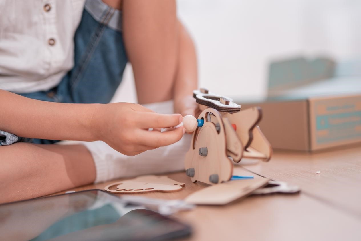 A child's hand carefully places a wooden bead onto a wooden toy, focusing intently on the task. The scene is a close-up, emphasizing the detail of the construction and the child's concentration.