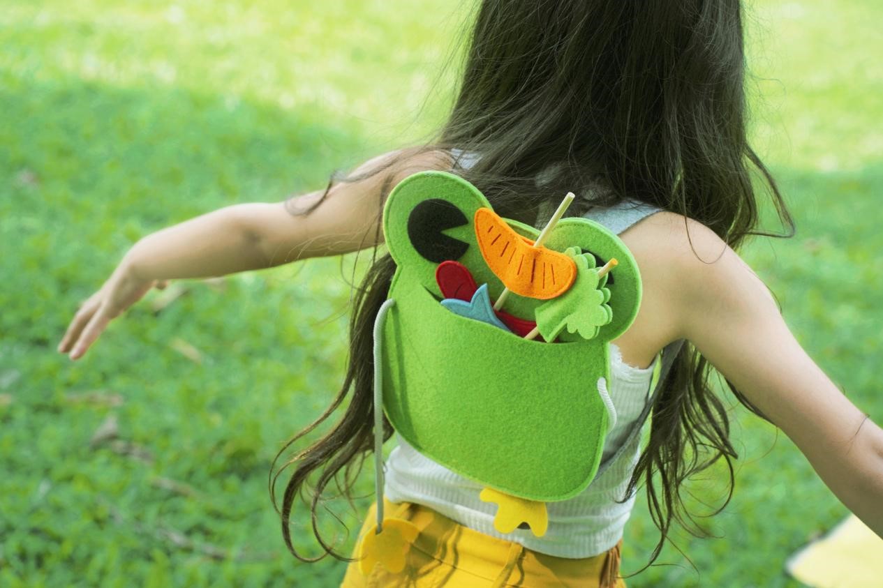 A young girl with long dark hair, wearing a white tank top and yellow shorts, walks through a grassy field, her arms outstretched, as if she is flying. She is wearing a green felt backpack shaped like a frog, with a variety of felt food items sticking out of the top, including a carrot, a fish, and a piece of lettuce. The scene is playful and imaginative, capturing the joy of childhood and the power of pretend play