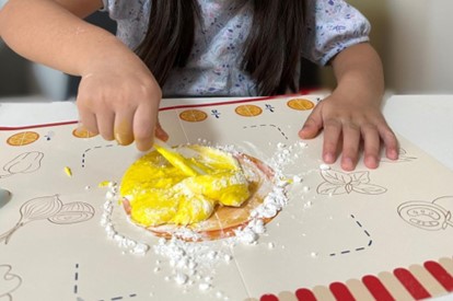 A child's hand carefully mixes yellow playdough with a yellow spatula on a white surface. The playdough is surrounded by white powder, creating a contrasting and playful scene. The child's hand is small and delicate, highlighting the joy of exploration and creativity in making pizza dough.