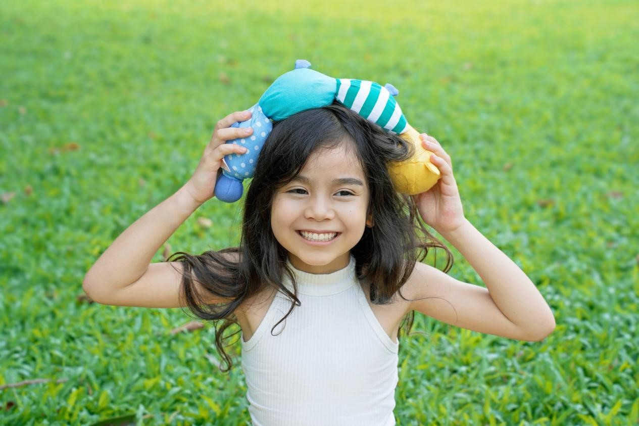 A young girl with long dark hair is holding a stuffed toy above her head, smiling brightly at the camera. She is wearing a white tank top and is surrounded by lush green grass. The scene is full of joy and innocence, capturing the simple pleasures of childhood.