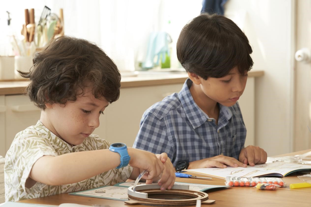 Two young boys are sitting at a table, engrossed in a craft project. The boy on the left is concentrating intently on his work, while the boy on the right is looking at his instructions. The scene is peaceful and heartwarming, capturing the joy of learning and the beauty of childhood.