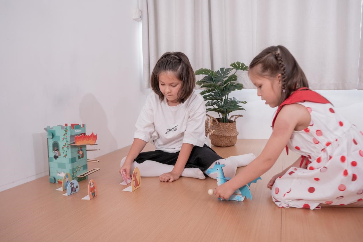 Two young girls are playing a game with a wooden castle and a dragon toy. They are both focused on the game, their faces lit up with excitement. The scene is playful and imaginative, capturing the joy of childhood and the power of pretend play.