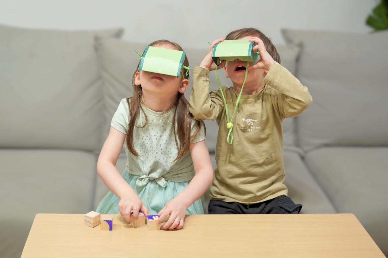 A young girl and boy are sitting at a table, wearing cardboard mirror periscope headsets. The girl is playing with wooden blocks, while the boy is looking around in awe. The scene is playful and imaginative, capturing the joy of childhood and the power of pretend play.