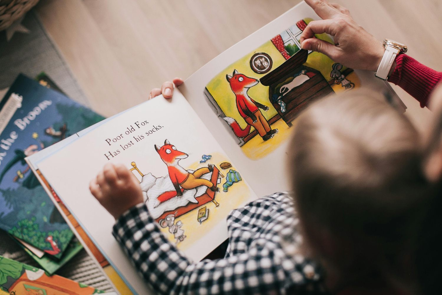 An overhead view of an adult and a child reading a children's book together. The book is open to a page with illustrations of a fox and the text Poor old Fox Has lost his socks. The adult's hand is lifting a flap on the page, while the child, wearing a black and white checkered shirt, looks on. Other books are visible on the floor around them.