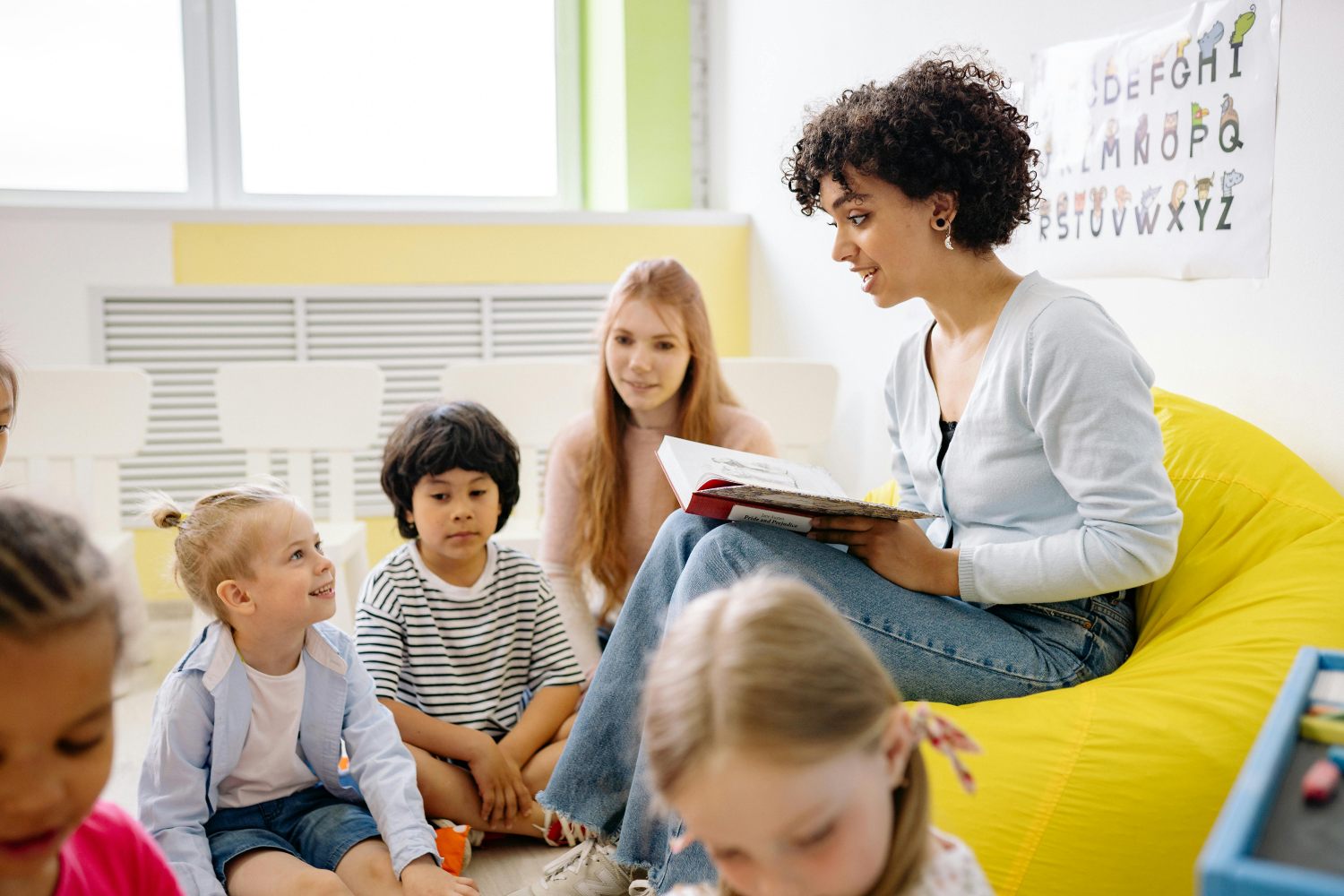 A female teacher with curly hair sits on a large yellow beanbag chair in a classroom, reading a book to a diverse group of young children. The children are sitting on the floor, listening attentively. Another teacher is visible sitting behind the children, and an alphabet poster hangs on the wall in the background.